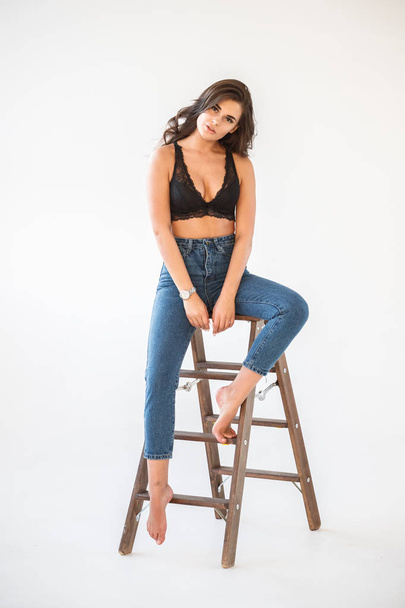 Studio portrait of beautiful curly brunette girl wearing blue jeans and sexual black underwear leaning on wooden staircase, white wall background - Foto, imagen