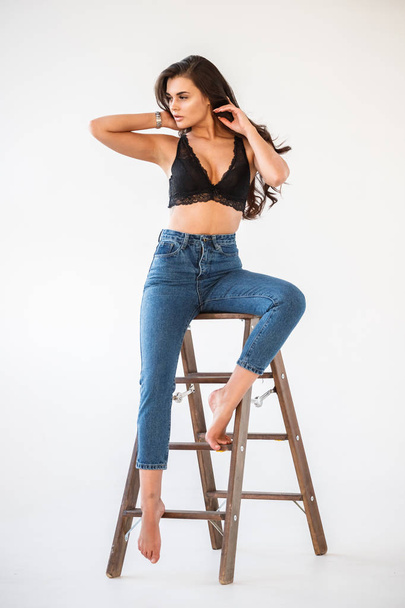 Studio portrait of beautiful curly brunette girl wearing blue jeans and sexual black underwear leaning on wooden staircase, white wall background - Photo, image