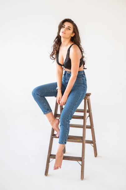 Studio portrait of beautiful curly brunette girl wearing blue jeans and sexual black underwear leaning on wooden staircase, white wall background - Fotó, kép