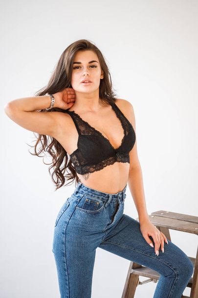 Studio portrait of beautiful curly brunette girl wearing blue jeans and sexual black underwear leaning on wooden staircase, white wall background - Foto, immagini