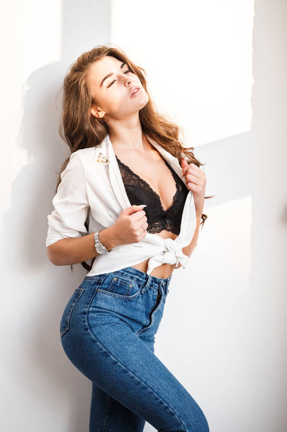 Studio portrait of beautiful curly brunette girl wearing blue jeans, white shirt and sexual black underwear standing on white wall background - Fotoğraf, Görsel