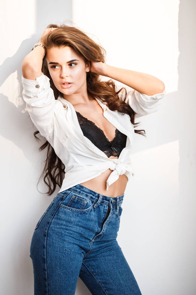 Studio portrait of beautiful curly brunette girl wearing blue jeans, white shirt and sexual black underwear standing on white wall background - 写真・画像