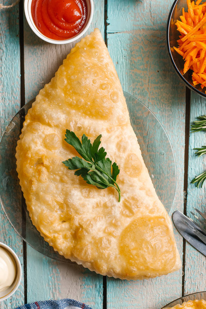 Closeup of cheburek decorated with parsley served on glass dish on blue wooden table with mayonnaise, ketcup and sliced carrot - Photo, Image