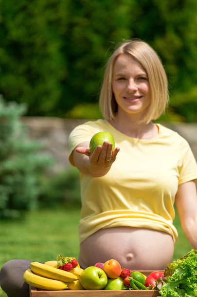 Pregnant woman sitting on grass and holding out apple in hand - Foto, immagini