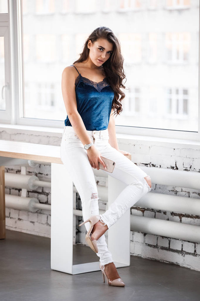 Portrait of young sexy brunette girl in blue lingerie and white pants sitting on table near window at studio  - Photo, Image