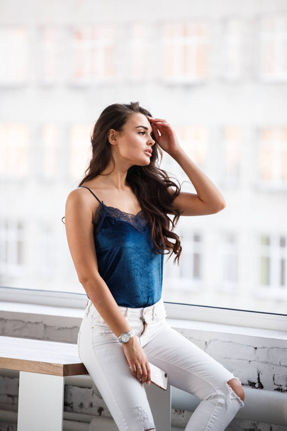 Portrait of young sexy brunette girl in blue lingerie and white pants sitting on table near window at studio  - Foto, Imagen