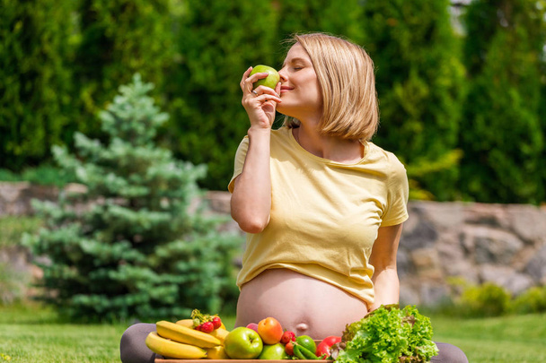 Pregnant woman sitting on grass and holding an apple in hand - Фото, зображення