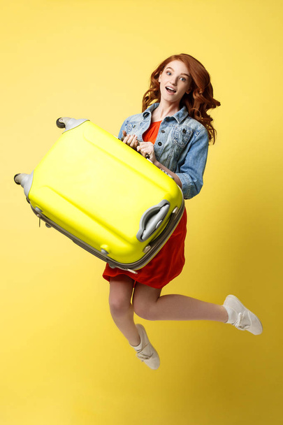 Travel and Lifestyle Concept: Portrait of a beautiful red hair fashion woman jumping and holding a bright green suitcase isolated over bright yellow background. - Φωτογραφία, εικόνα
