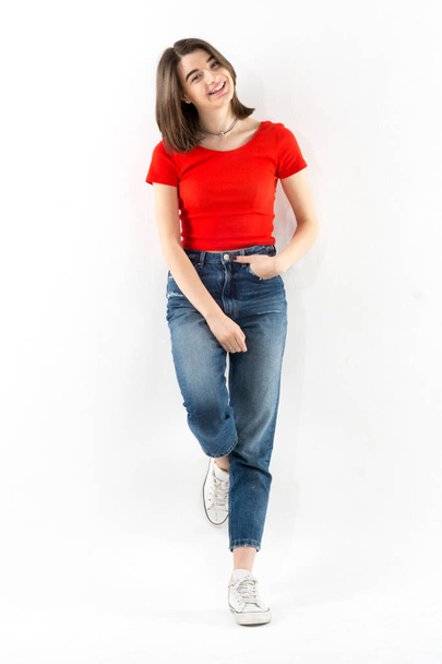 Beautiful young girl wearing red t-shirt and jeans leaning on a wall on white background, studio shoot, full-length portrait - 写真・画像