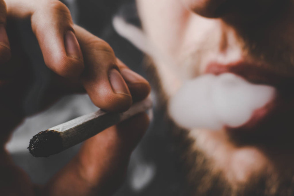 Ein Mann raucht Cannabiskraut, einen Joint und ein Feuerzeug in den Händen. Rauch auf schwarzem Hintergrund. Konzepte des medizinischen Marihuana-Konsums und der Legalisierung von Cannabis. - Foto, Bild