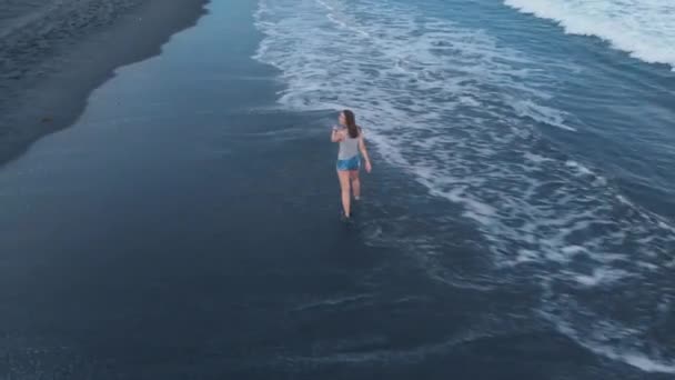 Filmato aereo della spiaggia oceanica con sabbia nera e onde al tramonto su Bali con bella felice giovane camminare, saltare, correre modello femminile in pantaloncini jeans e una maglietta grigia, riprese video 4K dal velivolo dall'alto, splendida vista, aria ki
 - Filmati, video