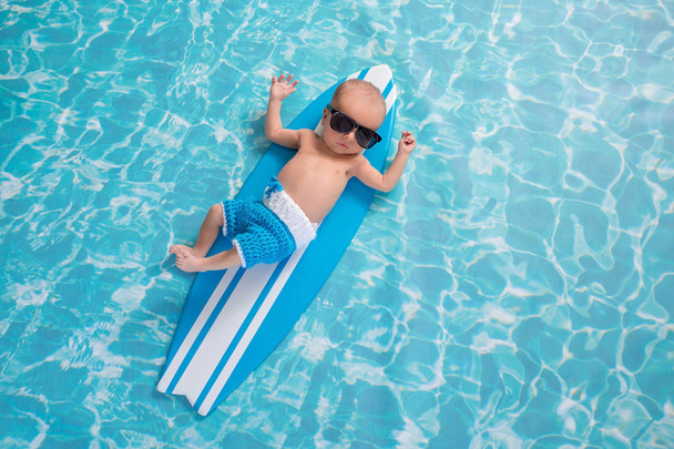One month old newborn baby boy lying on a tiny, light blue and white surfboard. He is wearing light blue, crocheted board shorts and sunglasses. - Photo, Image