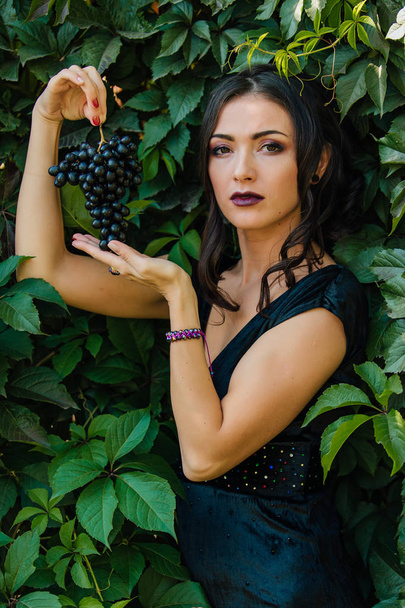 Portrait of young sexy brunette in black dress with black grapes. . - Fotoğraf, Görsel