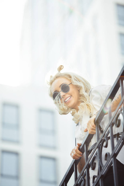 stylish woman with blond hair in sunglasses smiling holding on railing. - Foto, Imagen
