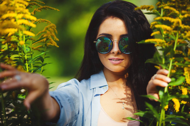 Horizontal portrait of beautiful woman in round sunglasses peeps out among yellow flowers. - Foto, Imagen