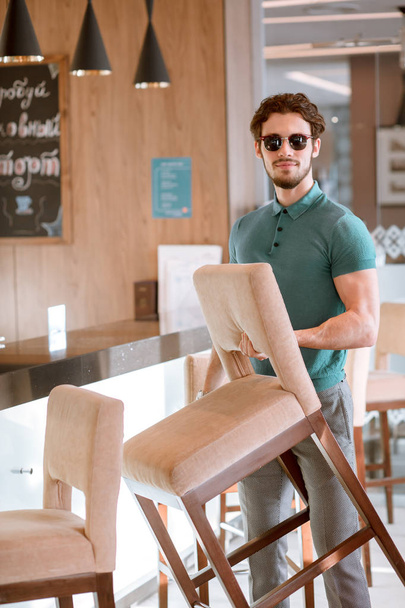 Portrait of handsome young bearded male lifting the chair at cafe. - Фото, зображення