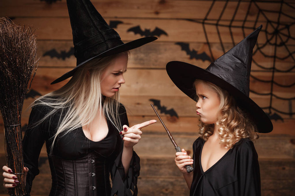 Halloween Concept - stressful witch mother teaching her daughter in witch costumes celebrating Halloween over bats and spider web on Wooden studio background. - Photo, Image