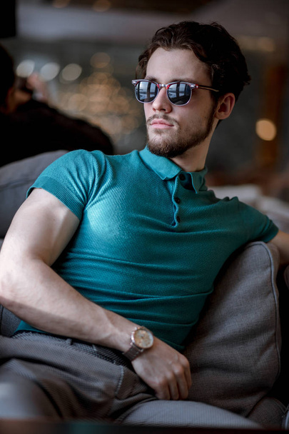 young Caucasian man in sunglasses sitting and ponder in armed chair - Фото, изображение