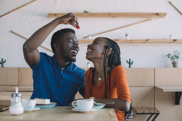 african american man feeding girlfriend with strawberry at table in cafe - 写真・画像