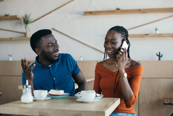 retrato de la sonriente mujer afroamericana hablando en smartphone con novio cerca en la cafetería
 - Foto, imagen