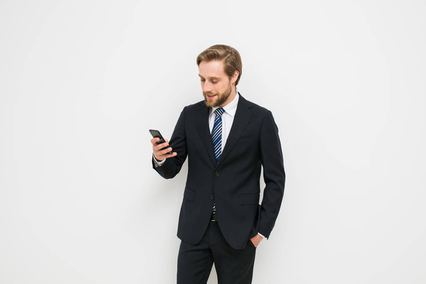 a cheerful businessman talking through phone messages with colleagues or family while at work, beautiful bearded and blond hair in elegant costume leaning against a white wall - Foto, immagini