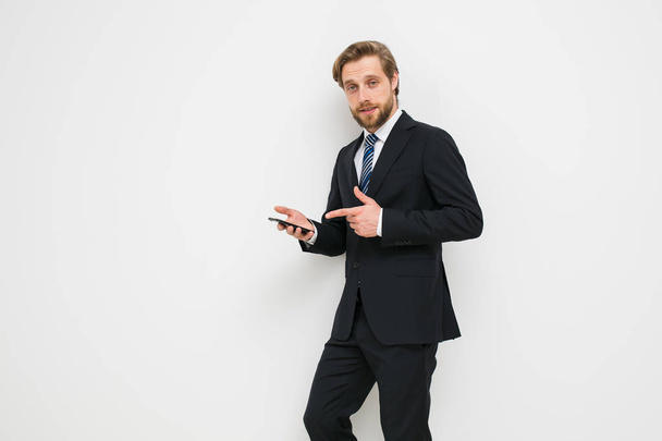 smart-looking guy pointing to his or her phone and what to do with it, or the application on the phone, relaxed in elegant costume on a white wall - Foto, imagen