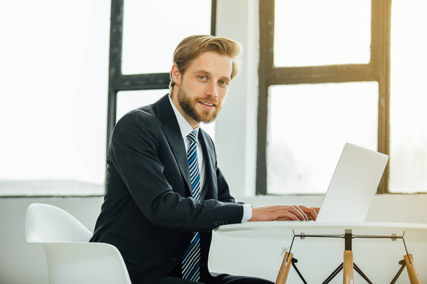 elegant man in suit, working on laptop in his office or window, looking at the camera with confidence - Photo, Image