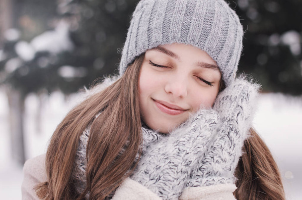 Οριζόντια πορτρέτο ενός νεαρού κοριτσιού σε γκρι πλεκτό καπέλο, ζεστά πλεκτά γάντια και μπεζ παλτό. Εφηβικό κορίτσι με τα μάτια κλειστά κρατά τα χέρια κοντά στο πρόσωπό. Θετικά vibes. Χειμερινής περιόδου - Φωτογραφία, εικόνα