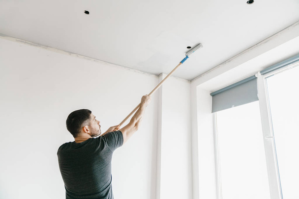 майстер у сірій футболці фарбує стелю валиком у сірий колір у своїй квартирі. Ремонт і будівництво
. - Фото, зображення