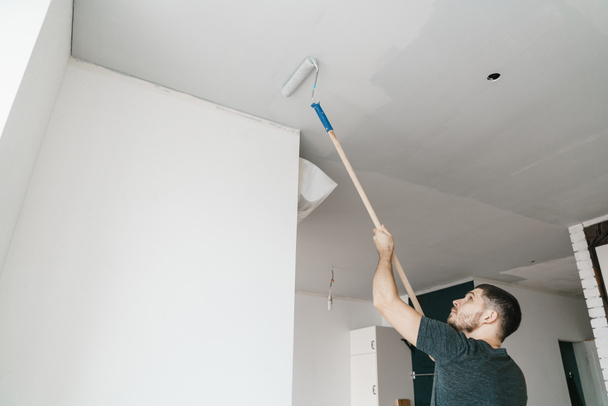 el hombre pinta el techo en un rodillo de color gris en casa. Reparación y renovación del apartamento
. - Foto, Imagen