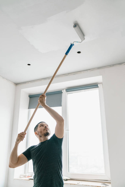 un hombre con un rodillo en la mano sobre un palo largo pinta el techo de gris en la ventana. Reparación en una habitación luminosa
. - Foto, imagen