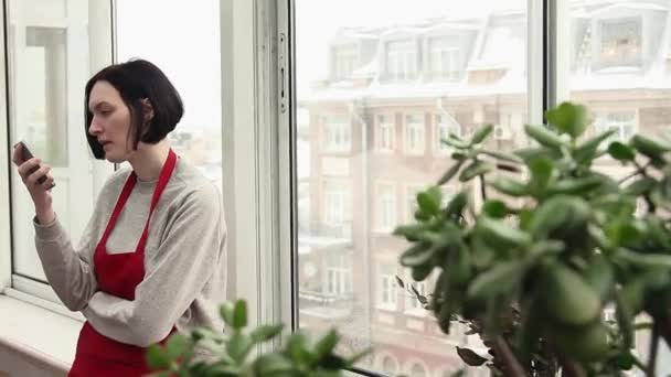 Young girl gardener in red apron chatting online using smartphone in greenhouse - Filmagem, Vídeo