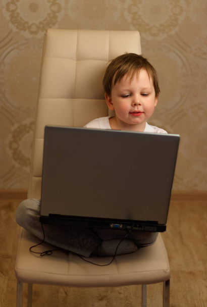 the boy is sitting on a chair and is holding a laptop - Фото, изображение