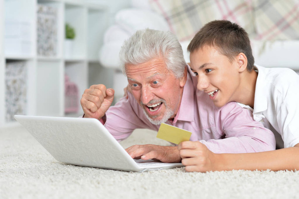 Nagyapa és unokája, feküdt a padlón, és használ a laptop, unokája holding hitelkártyát, online vásárlás koncepció - Fotó, kép