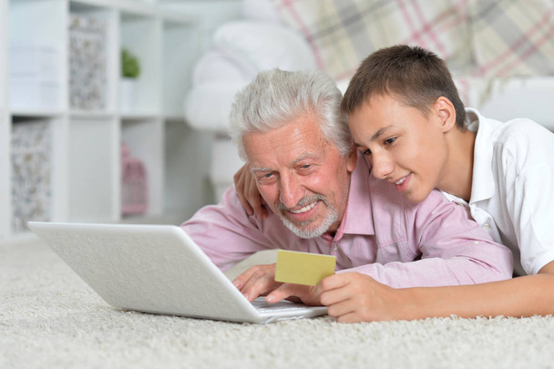 avô e neto deitado no chão e usando laptop, neto segurando cartão de crédito, conceito de compras online
 - Foto, Imagem