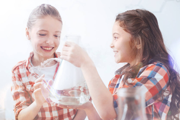 Excited schoolgirls grinning widely while holding flask in school lab - Photo, image