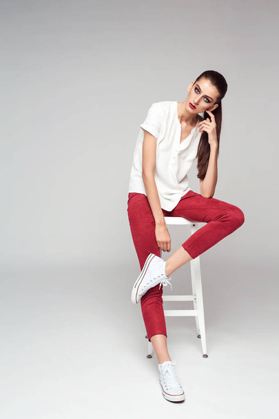 portrait of beautiful brunette girl in stylish clothes posing sitting on white chair, full length  - 写真・画像