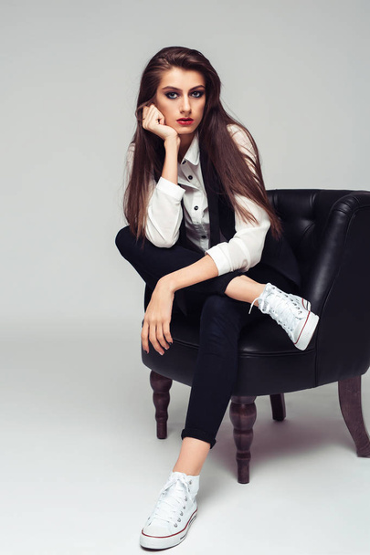 Young beautiful woman posing sitting on arm-chair in stylish clothes and keds, full length - Photo, Image