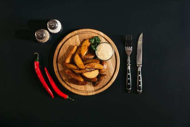 leckere Ofenkartoffeln mit Sauce auf Holzbrett, Gewürzen, Chilischoten und Gabel mit Messer auf schwarz   - Foto, Bild