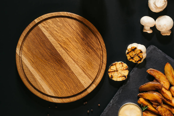 Blick von oben auf leeres Holzbrett, gegrillten Knoblauch, Pilze und Ofenkartoffeln mit Sauce auf Schwarz - Foto, Bild