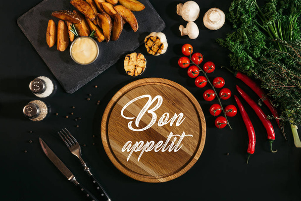 tablero de madera con inscripción bon appetit, papas al horno y verduras en negro
 - Foto, Imagen