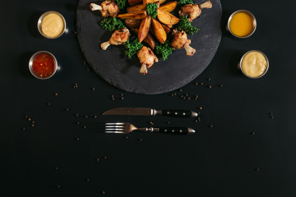 top view of delicious baked potatoes with fried chicken, various sauces and fork with knife on black - Foto, afbeelding