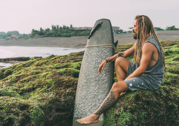 Tanned young surfer with tattoos and dreadlocks with his surfboard, sitting on the green mossy rocks near the ocean in Indonesia, Bali, Canggu, Batu Bolong beach and chilling before surfing. - 写真・画像