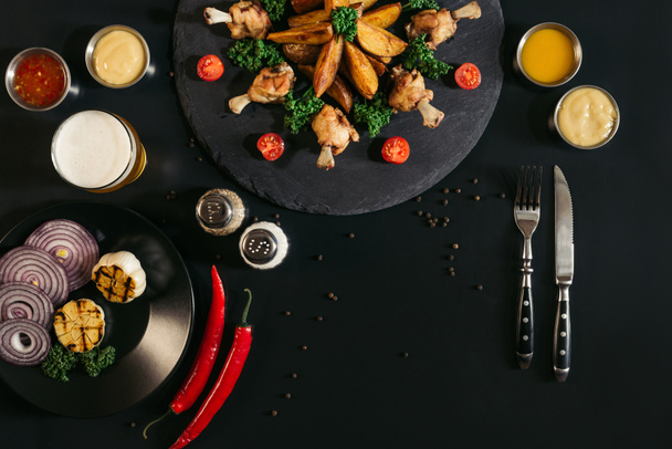 delicious roasted potatoes with chicken wings, sauces, vegetables and glass of beer on black  - Fotoğraf, Görsel