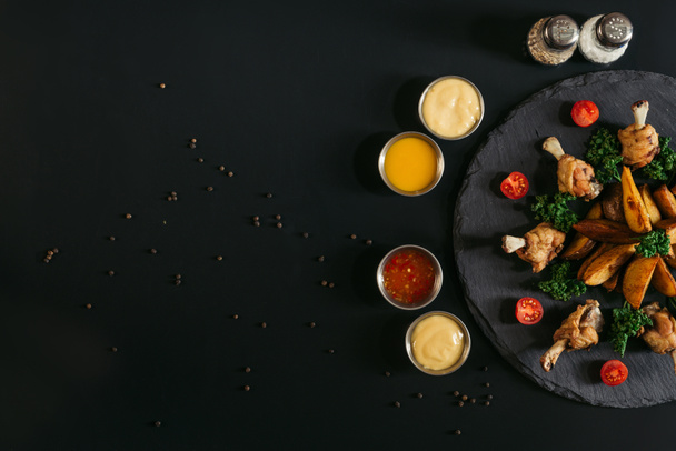 various sauces, spices and delicious baked potatoes with roasted chicken wings on slate board on black  - Fotografie, Obrázek