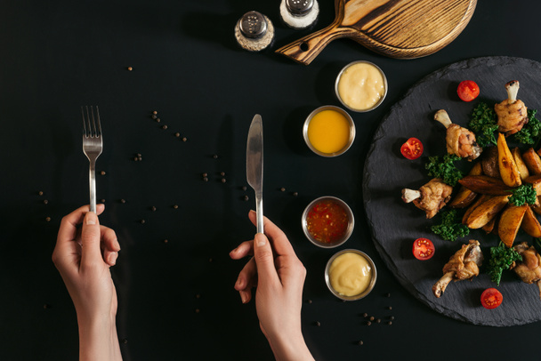 vue partielle du dessus de la personne tenant fourchette et couteau et délicieuses pommes de terre cuites au four avec poulet rôti et sauces sur noir
 - Photo, image