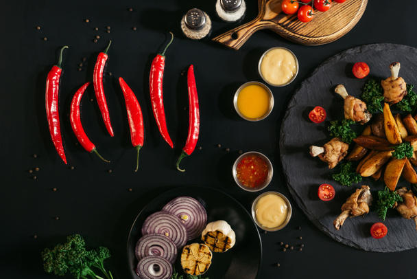 top view of red peppers, sauces, vegetables and tasty baked potatoes with roasted chicken on black - Fotoğraf, Görsel