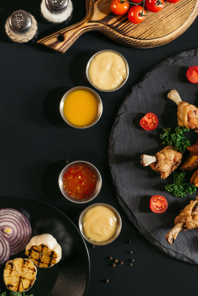 top view of tasty roasted chicken wings with various sauces and vegetables on black - Zdjęcie, obraz