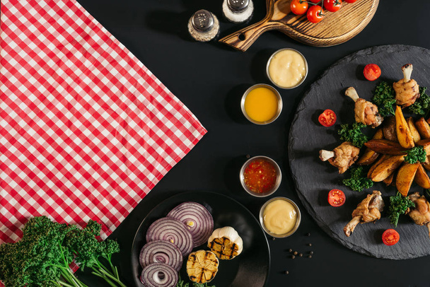 top view of checkered napkin, vegetables, sauces and delicious roasted potatoes with chicken on black  - Fotografie, Obrázek