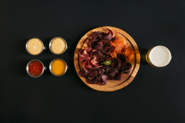 top view of delicious sliced assorted meat on wooden board with glass of beer and sauces on black - Fotoğraf, Görsel